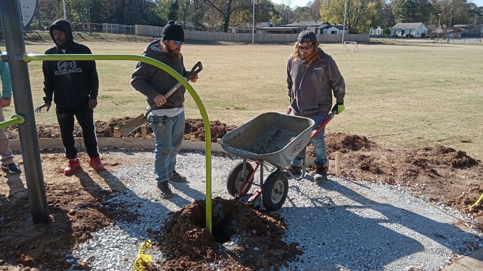 Parks workers about to pour concrete into hole that leg of exercise equipment is in